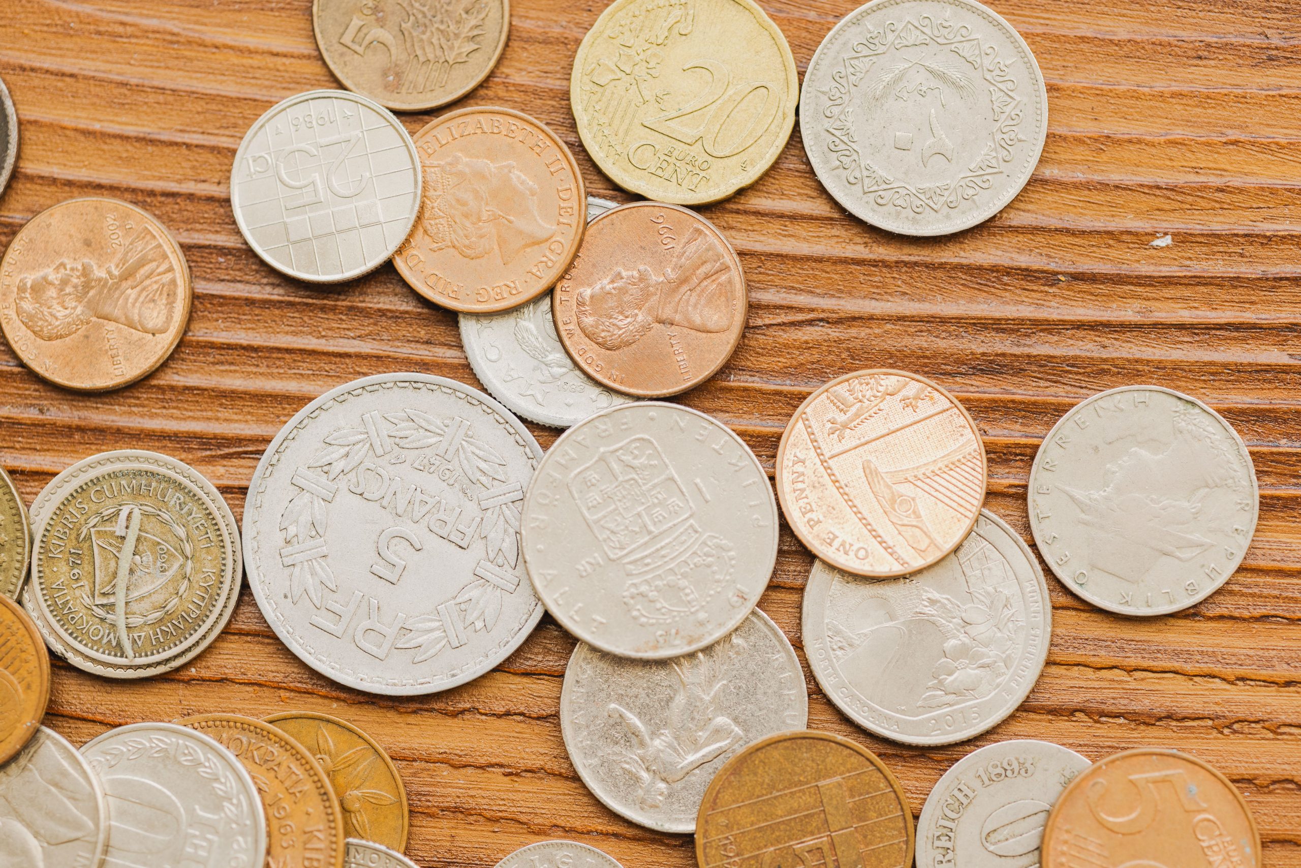 Mixed world and U.S. coins scattered across a wooden surface, including pennies and silver-colored foreign coins.